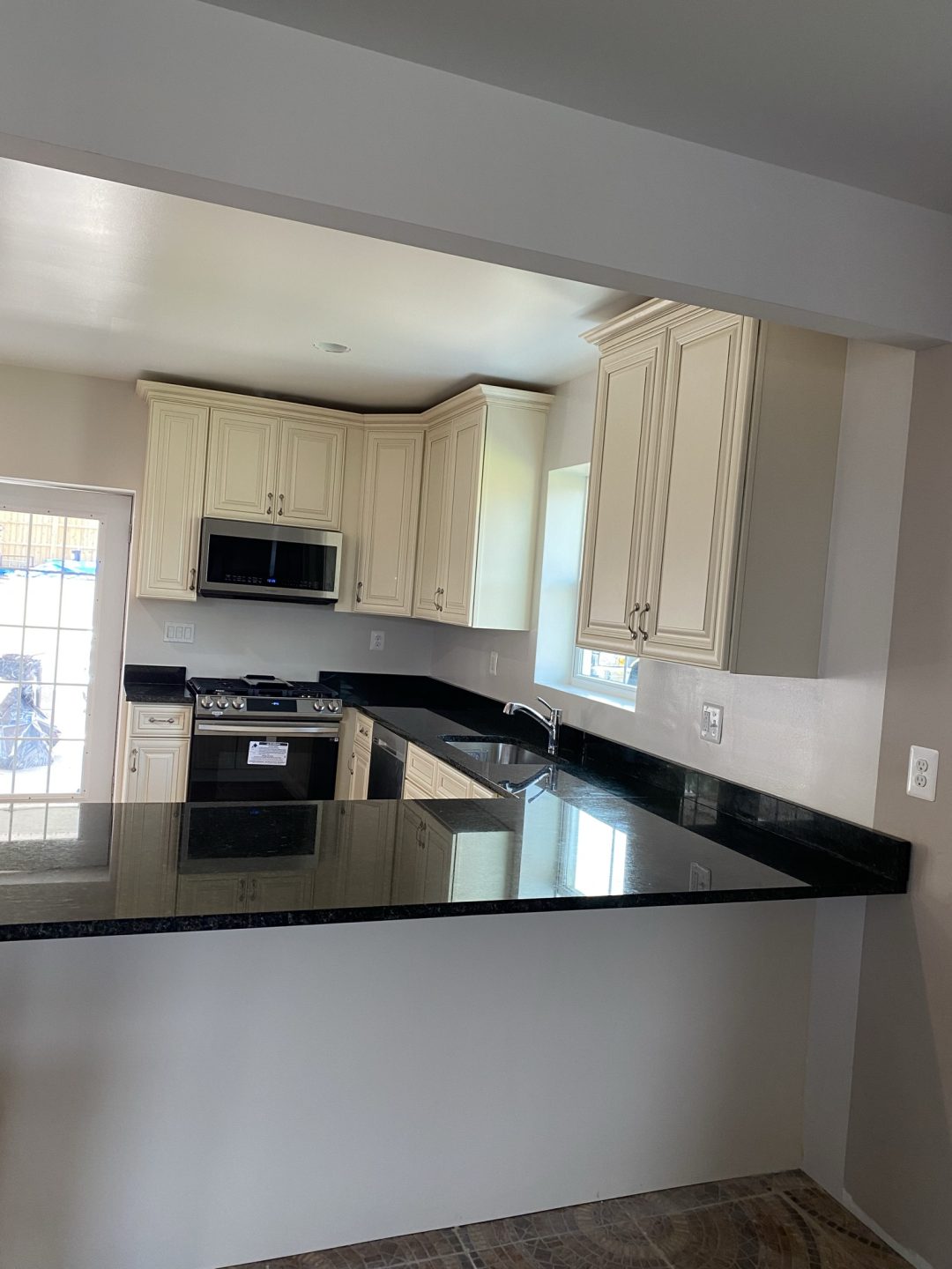 Modern kitchen with cream cabinets, black countertops, and stainless steel appliances, seen over a breakfast bar.