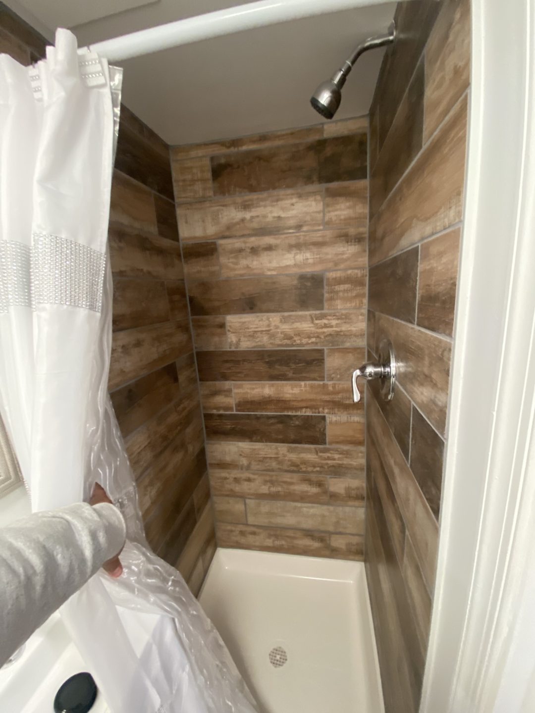 A person opens a shower curtain, revealing a shower with brown wood-look tiles and a metal showerhead.
