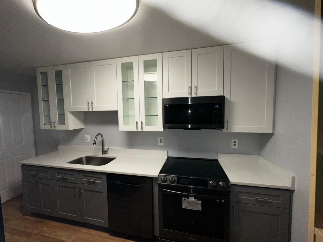 Modern kitchen with white upper cabinets, gray lower cabinets, stove, microwave, and a sink under bright lighting.
