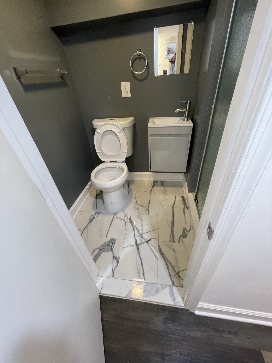 Small bathroom with gray walls, marble tile floor, toilet, sink, towel bar, and a mirror above the sink.