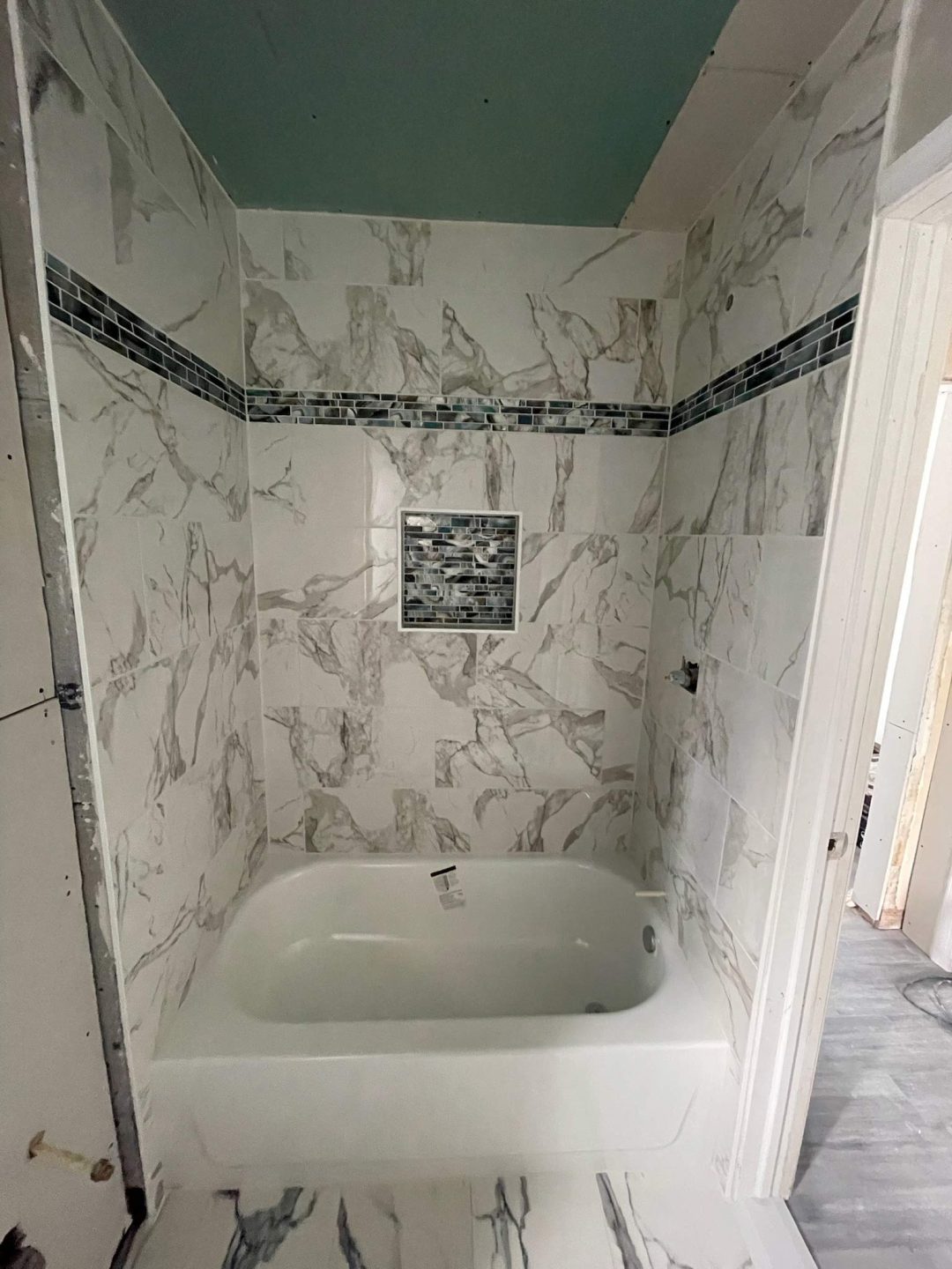 Small bathroom with a white bathtub, marble-patterned tiles, and a mosaic tile accent strip and niche.