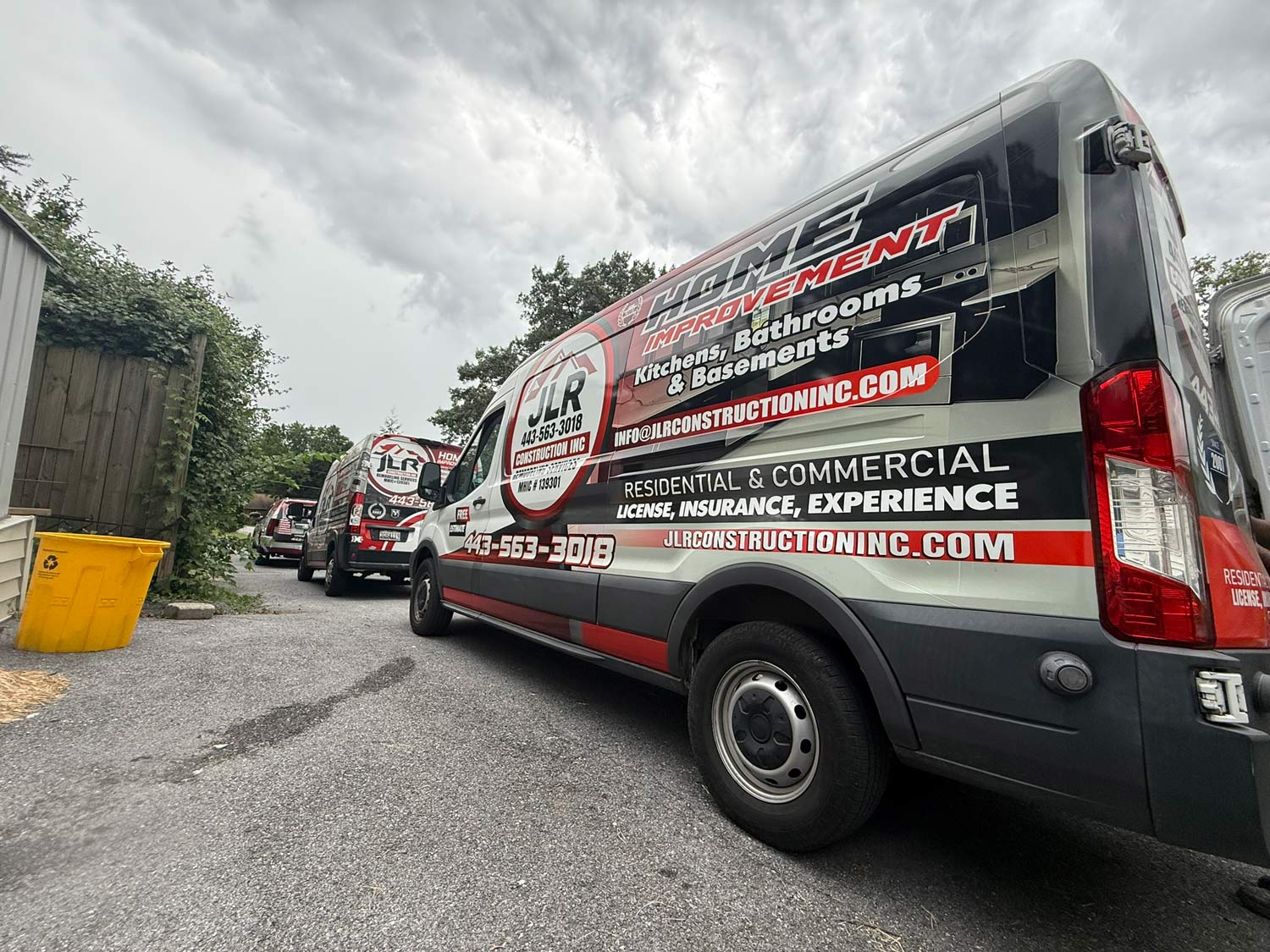 Three branded home improvement vans parked on a driveway under a cloudy sky, next to a yellow bin.