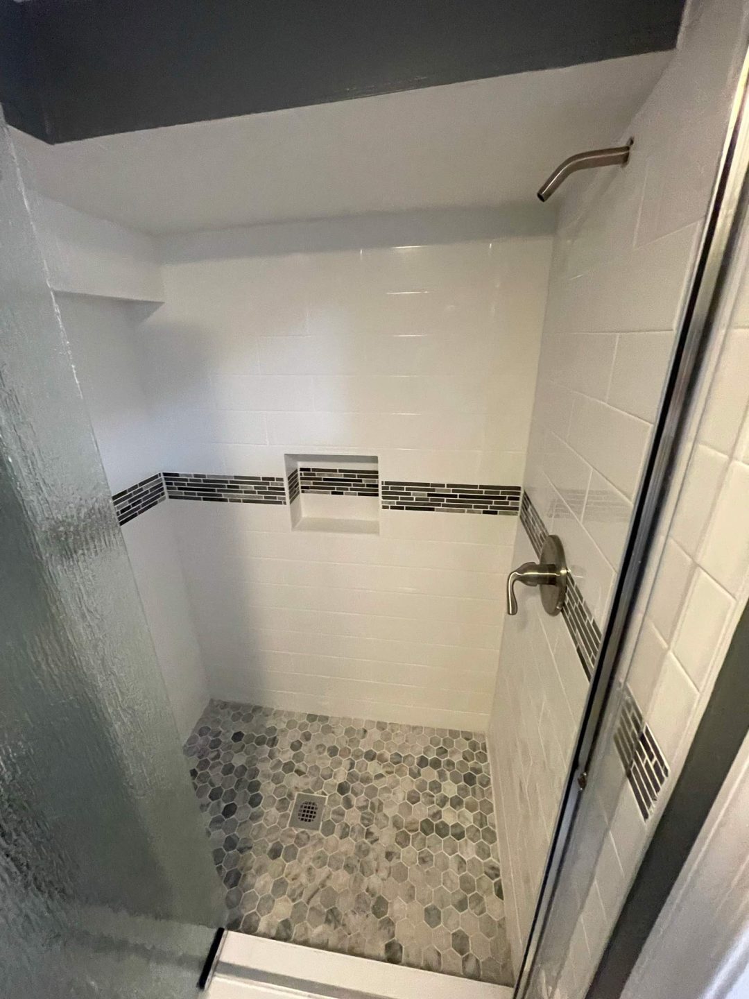 Modern walk-in shower with white tiles, mosaic accent strip, built-in shelf, and pebble stone floor.