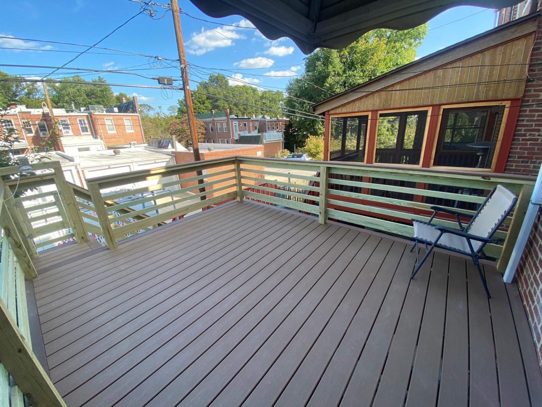 Spacious wooden deck with railings, a single chair, and views of nearby houses under a blue sky.
