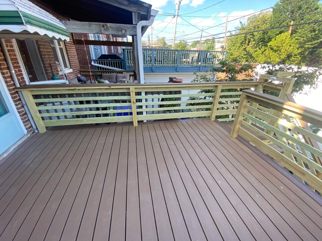 A wooden deck with a railing, overlooking neighboring buildings and trees on a sunny day.