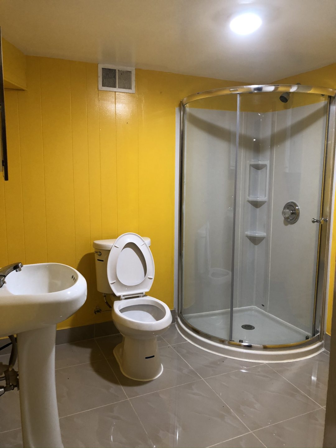 Bathroom with yellow walls, a toilet, pedestal sink, and a round glass shower enclosure under a ceiling light.