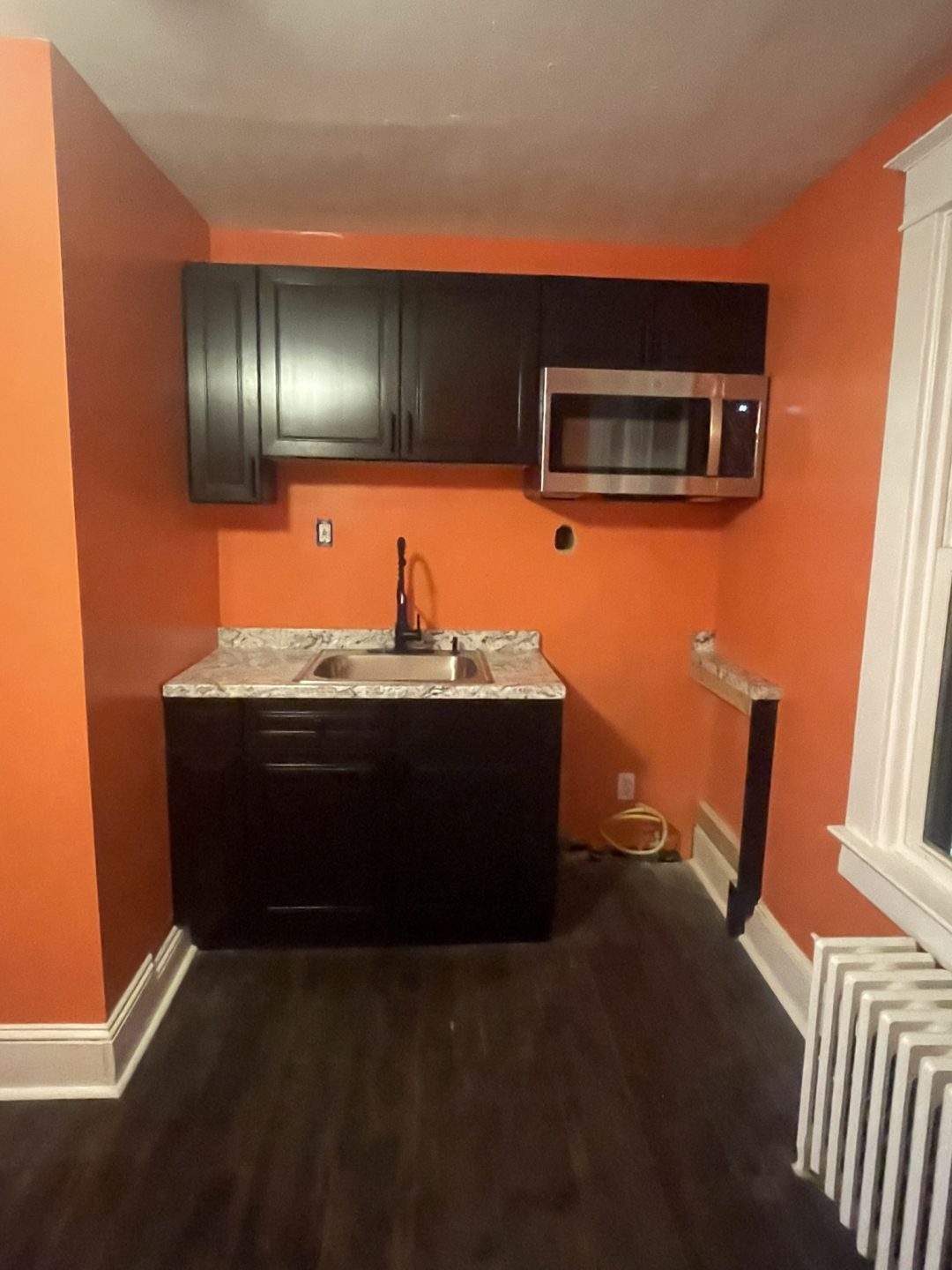 Small kitchen with orange walls, dark cabinets, a sink, microwave, and a marble-patterned countertop.