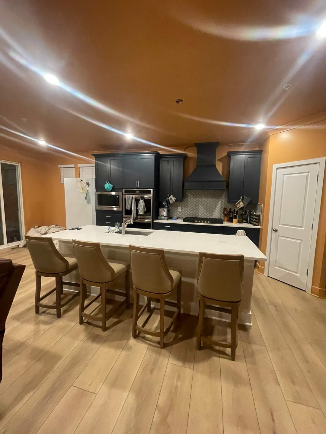 Modern kitchen with dark cabinets, a white island, four tan chairs, and wood floors under warm lighting.