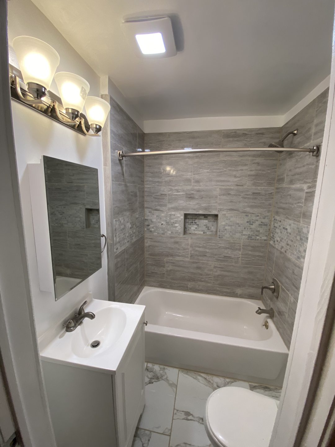 Small modern bathroom with gray tiled walls, white bathtub, vanity, mirror, toilet, and bright lighting.