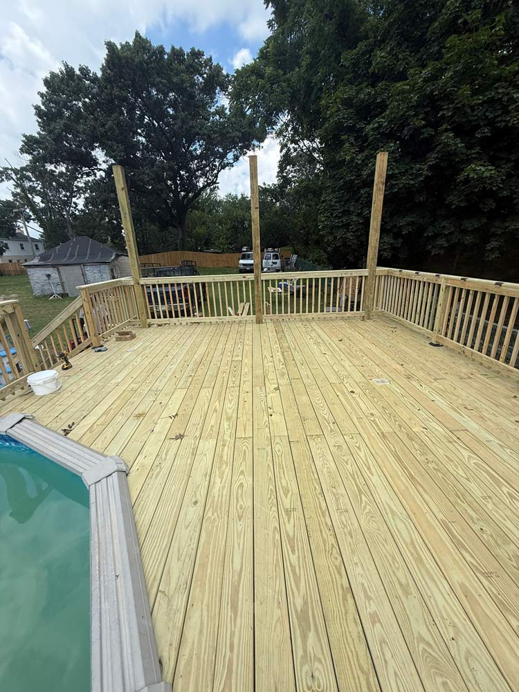 Newly built wooden deck with railing, beside a pool, and large trees in the background.