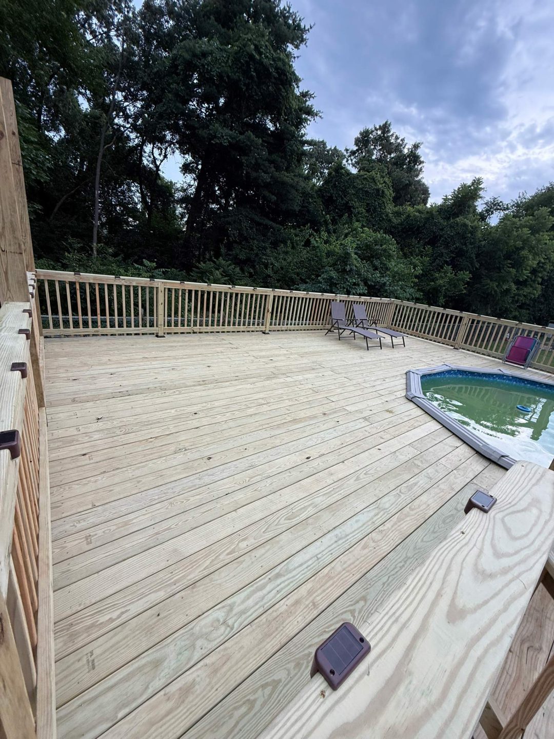 Spacious wooden deck with a pool, lounge chairs, and trees in the background under a cloudy sky.