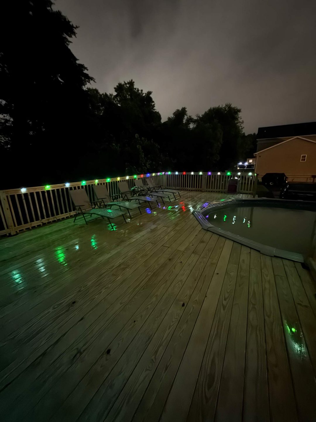 A wooden deck at night with colorful string lights reflecting on wet boards beside a covered pool.
