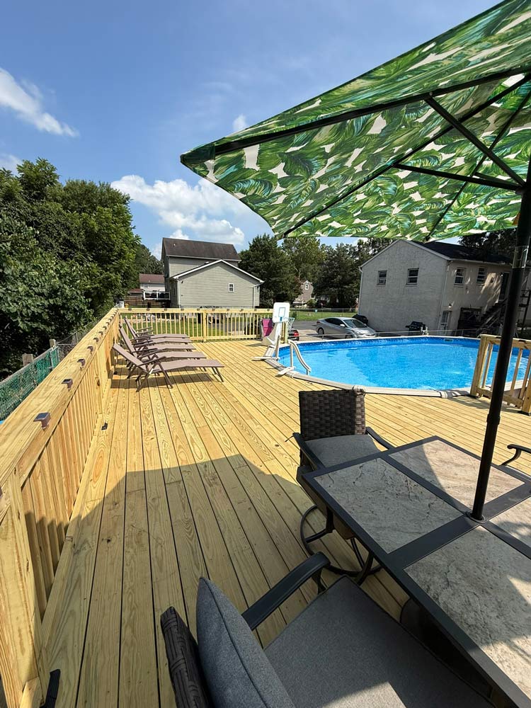 A sunny backyard deck with a pool, lounge chairs, and a table under a leaf-patterned umbrella.