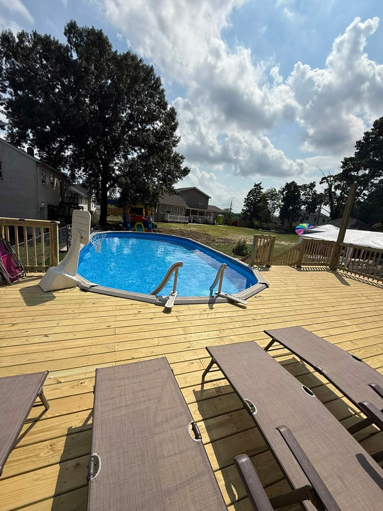Above-ground pool on a wooden deck with lounge chairs, surrounded by trees and houses under a partly cloudy sky.
