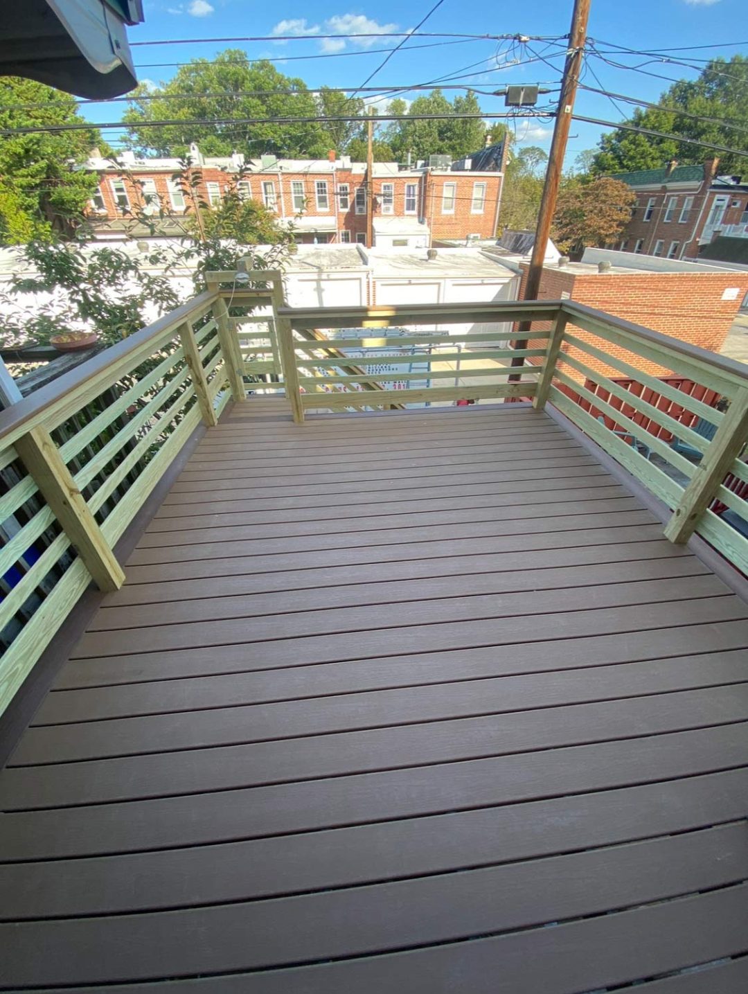 A wooden deck with railings overlooks a residential neighborhood with row houses and trees under a blue sky.