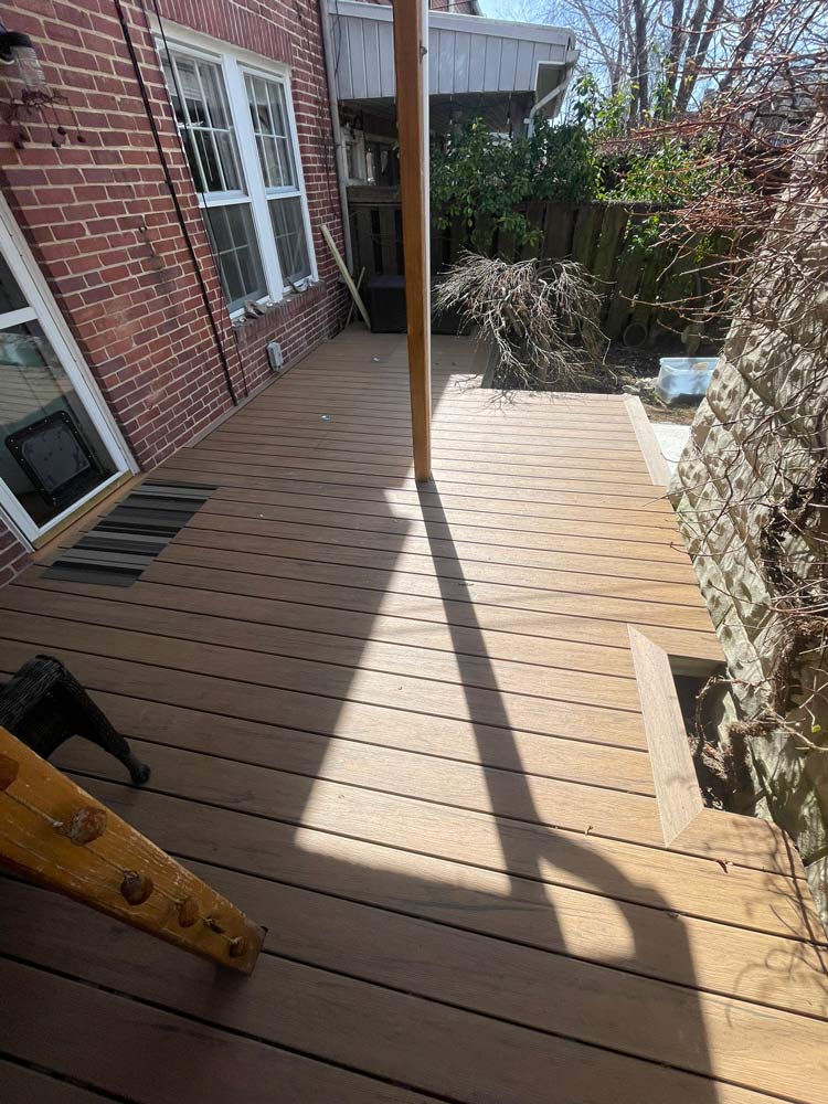 A wooden backyard deck with a central post, adjacent brick wall, and some plants along the edge.