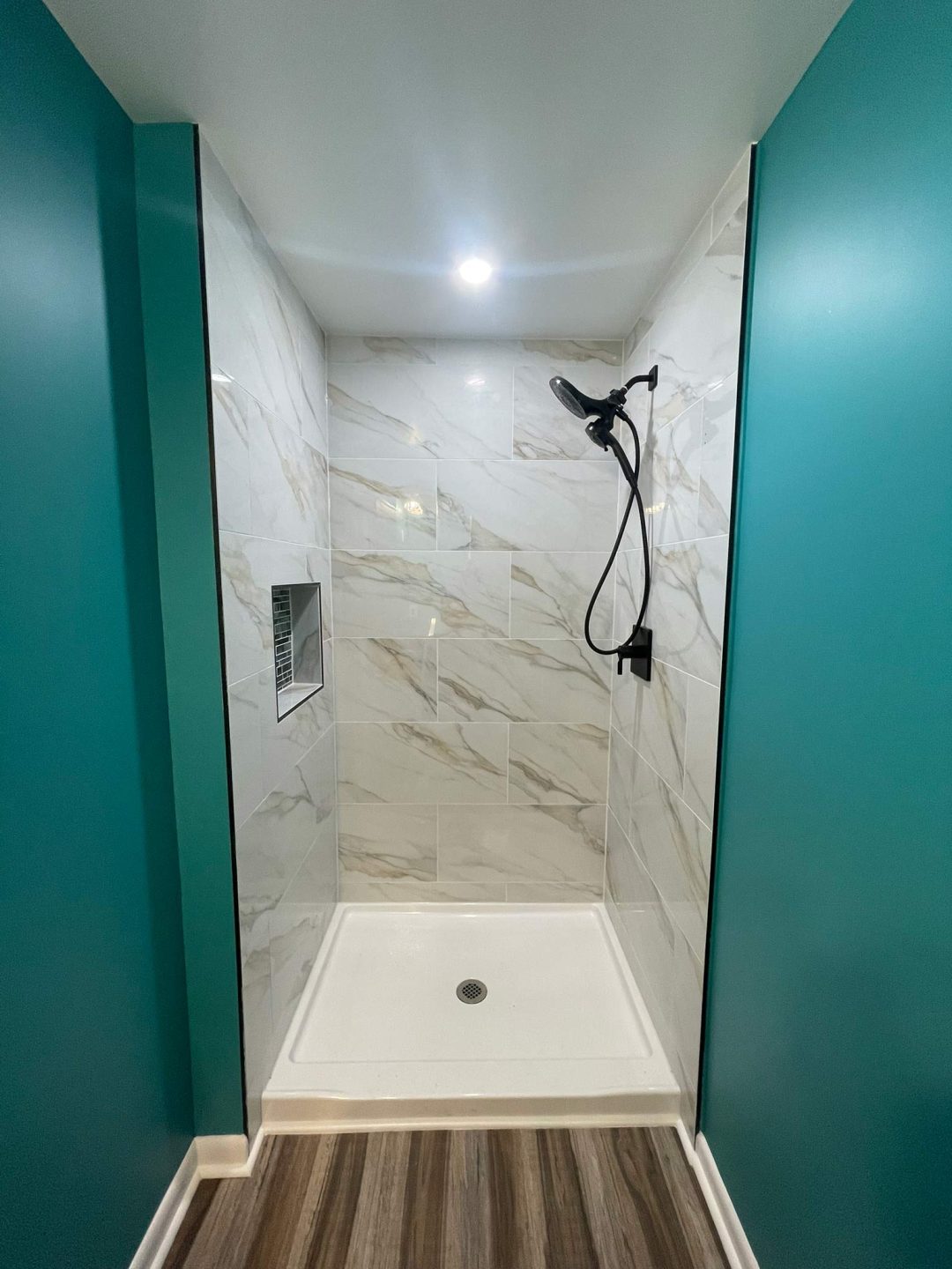 Modern shower with white marble tiles, teal walls, a black showerhead, and wood-look flooring.