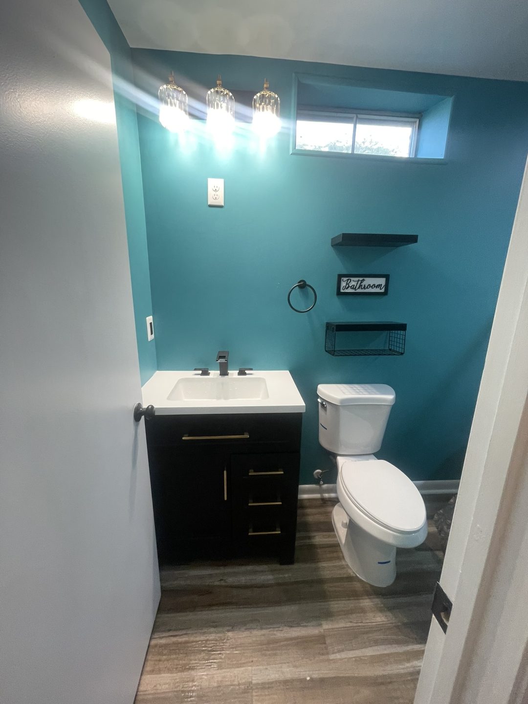 Small teal bathroom with a black vanity, toilet, wall shelf, and "Bathroom" sign; natural light above.