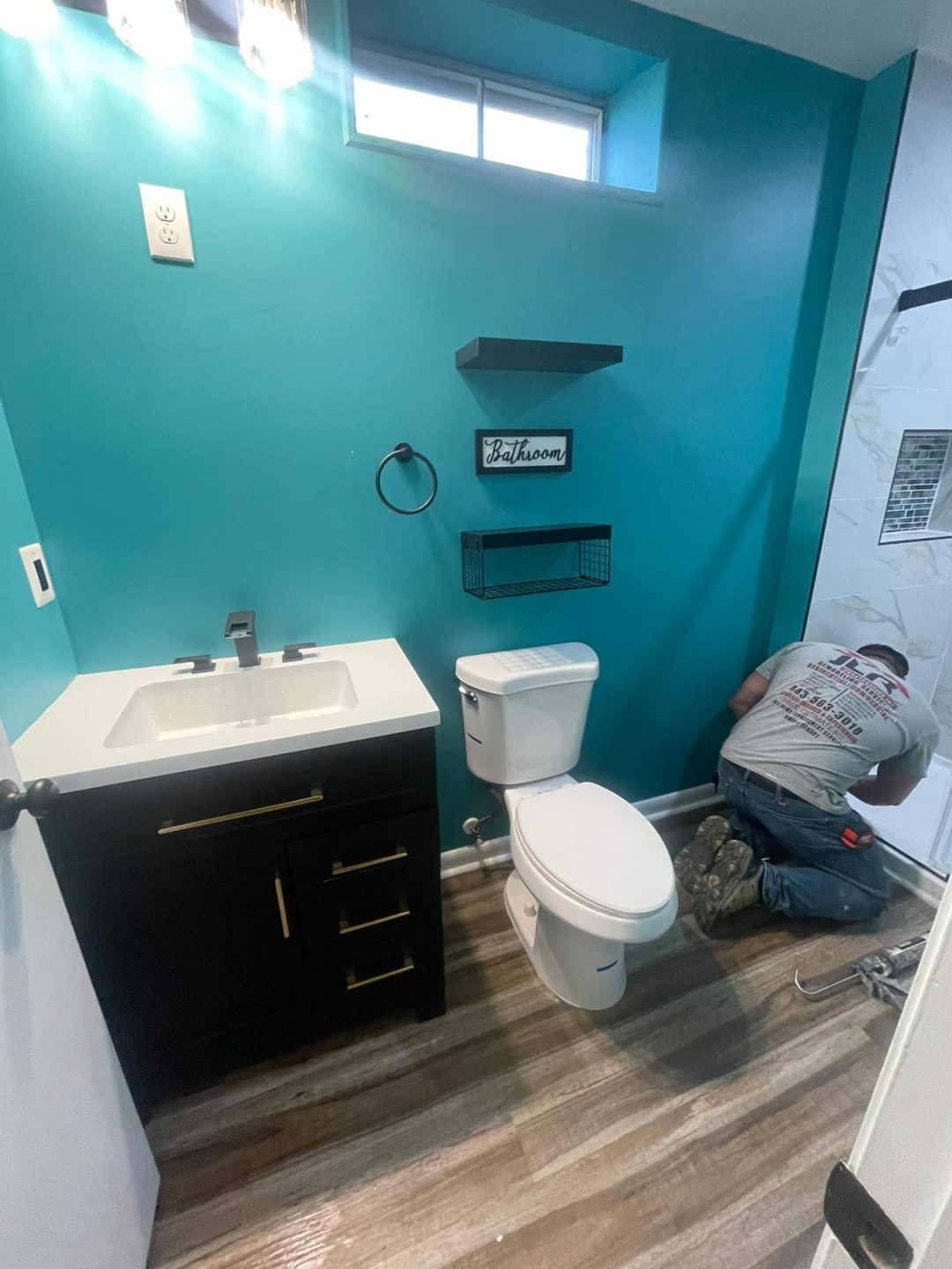 A person works on the floor of a teal bathroom with a vanity, toilet, and floating shelves.