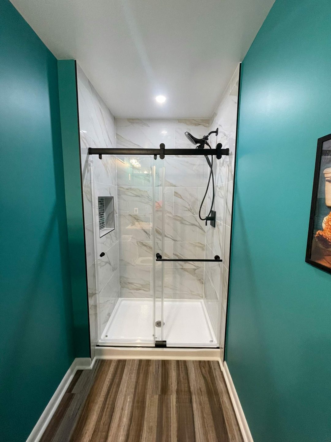 Modern shower with glass doors, white tile walls, black fixtures, teal walls, and wood-look flooring.