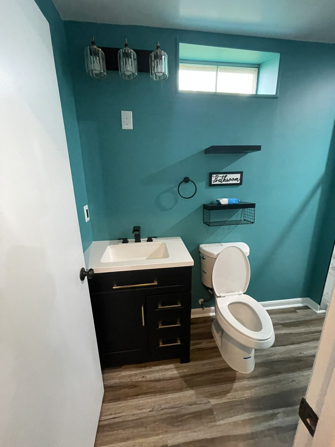 Small teal bathroom with a white sink, black vanity, toilet, and shelf with "Bathroom" sign above it.