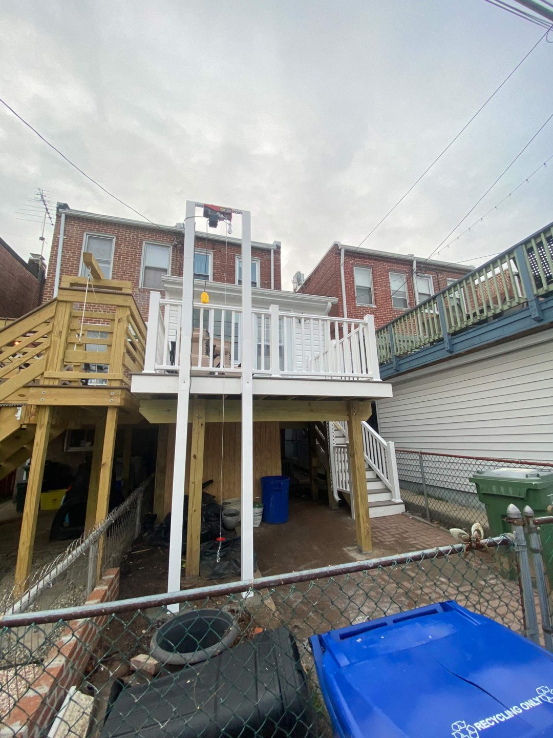 Two-story wooden and white-railed deck attached to a brick row house, with stairs and trash bins below.