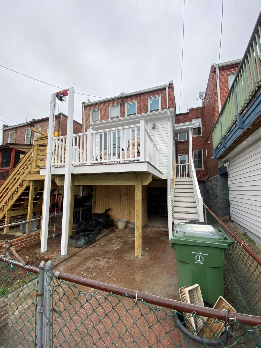 A raised white deck with stairs behind a brick house, over a concrete patio with garbage bins and potted plants.