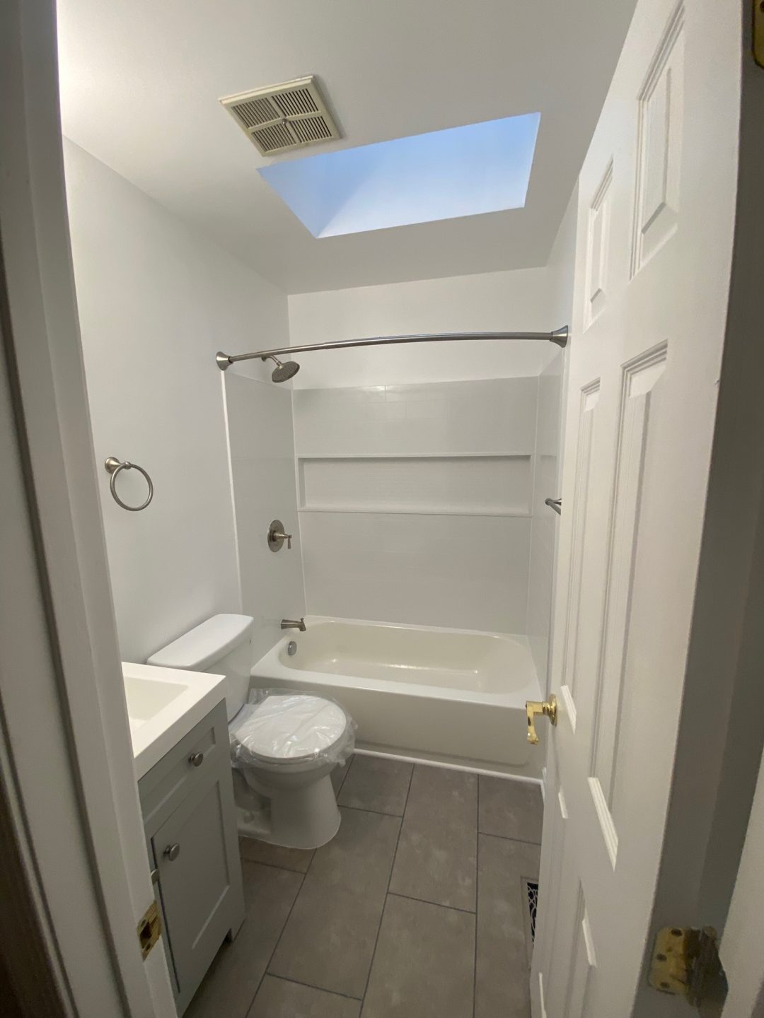 Small white bathroom with a tub, toilet, sink, tiled floor, and a skylight above.