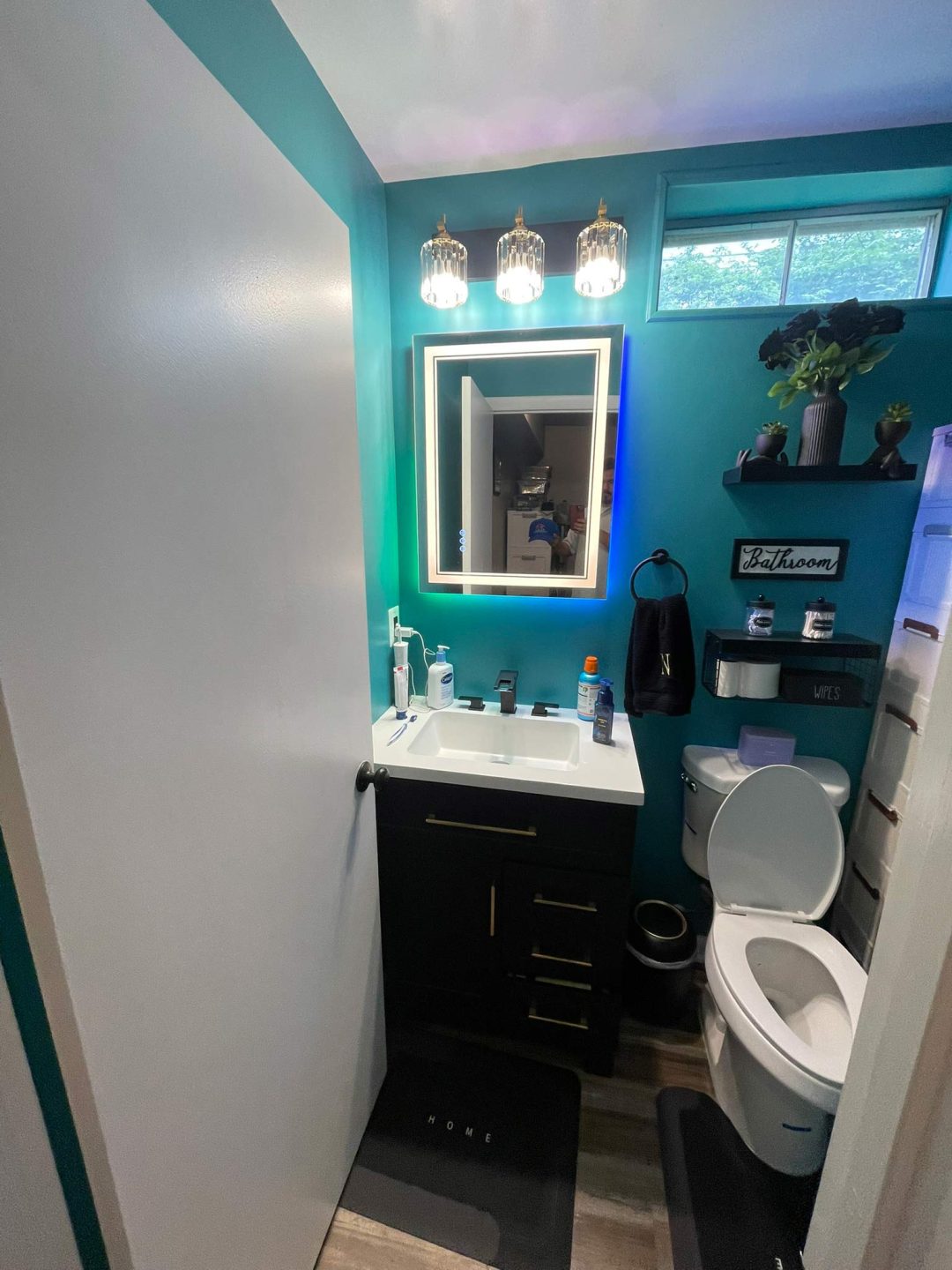 Small modern bathroom with teal walls, lighted mirror, black vanity, and toilet with seat open.