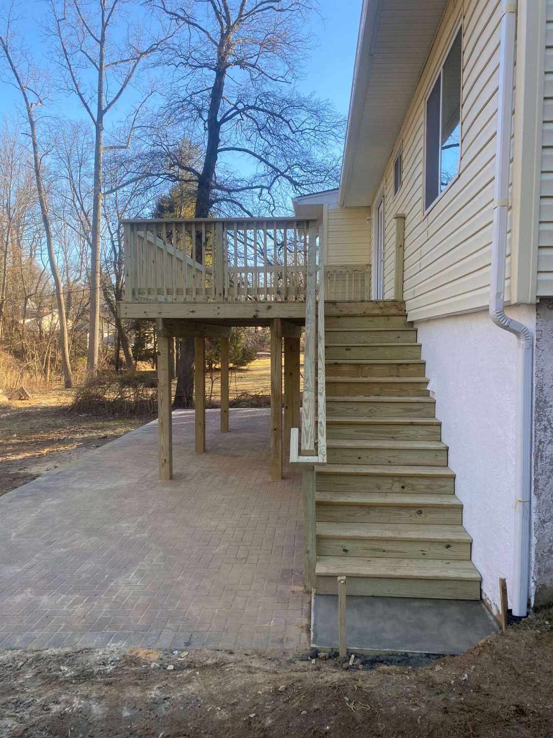 A wooden deck with stairs leads to a house, above a paved patio in a backyard.