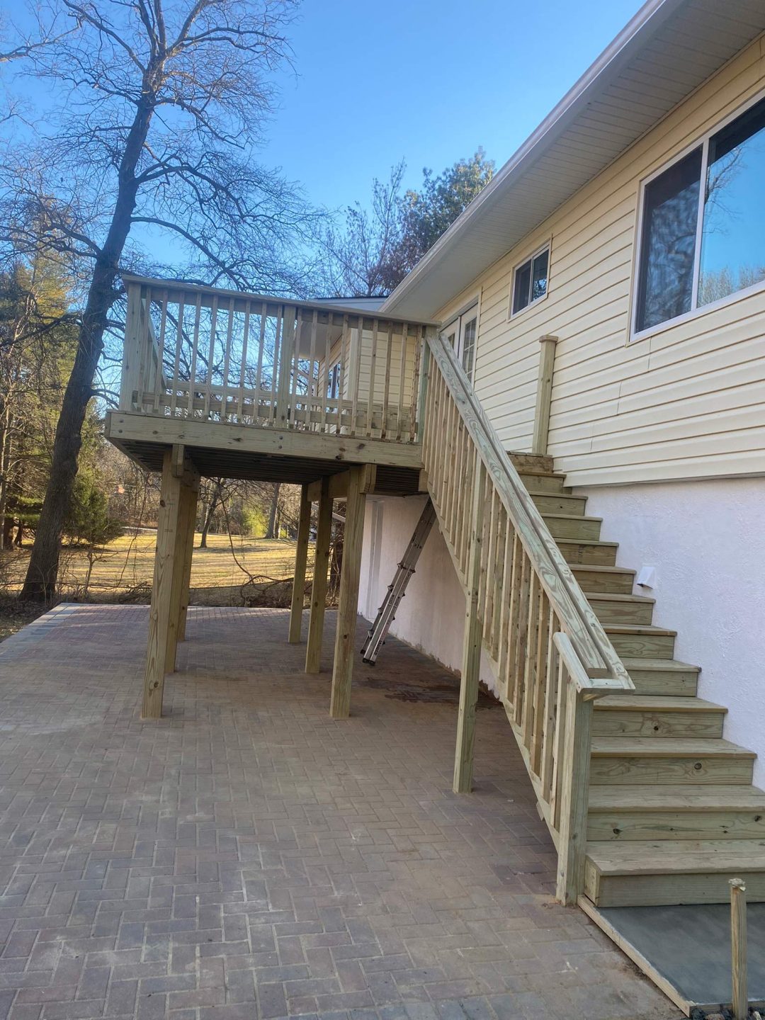 Wooden staircase and deck attached to a beige house, overlooking a brick patio and a grassy yard with trees.