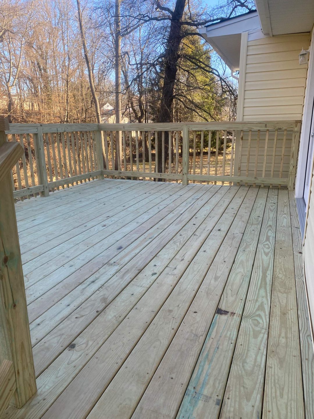 A wooden deck attached to a beige house overlooks a yard with leafless trees on a sunny day.