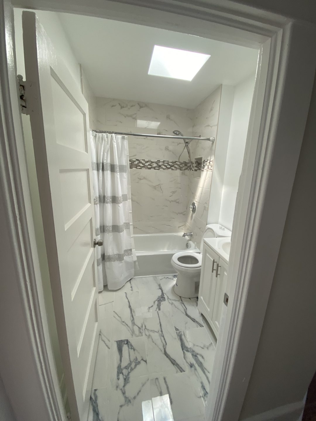 Bright bathroom with marble floor, bathtub with shower, toilet, white vanity, and a skylight above.