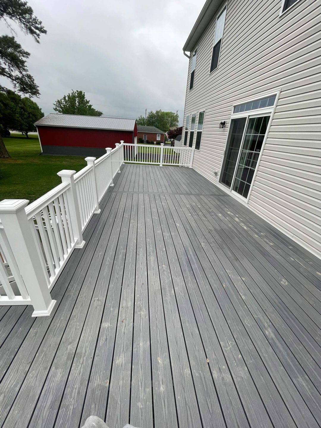 A gray wooden deck with white railings next to a house, overlooking a grassy yard and red barn.