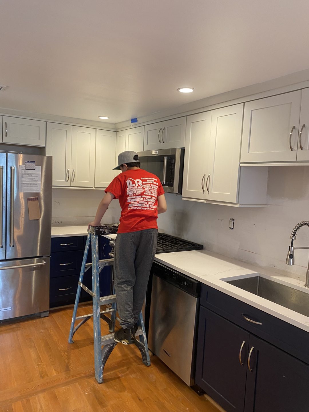 Person on a step ladder working on a kitchen cabinet above a stove in a modern kitchen.