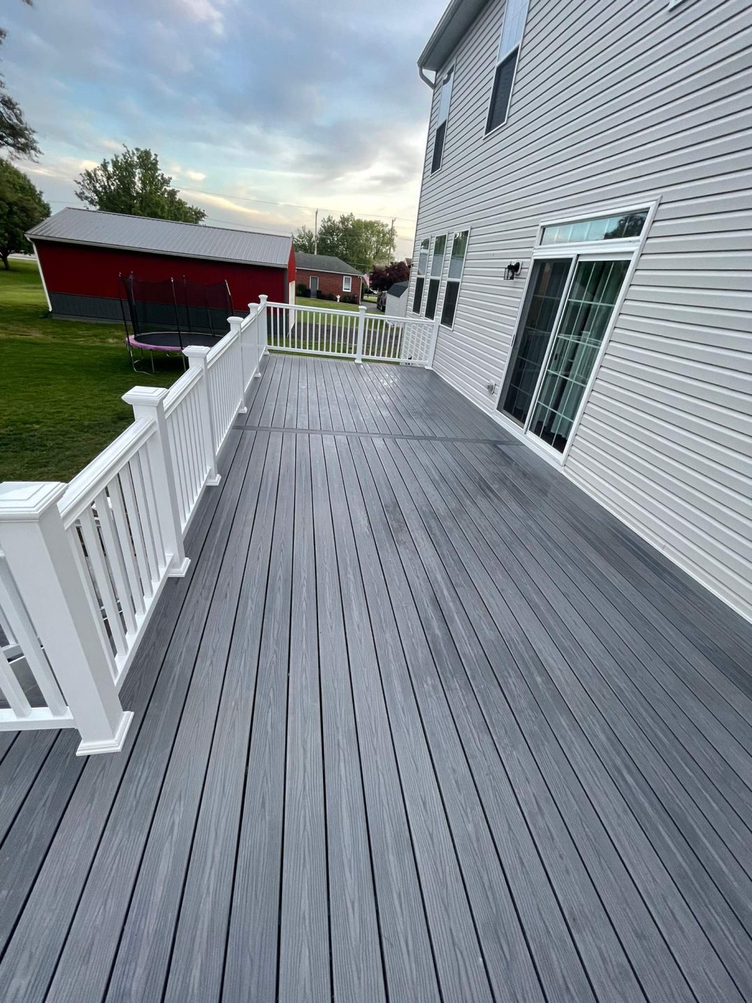 Wide gray deck with white railing attached to a white house, overlooking a grassy yard with a red shed.
