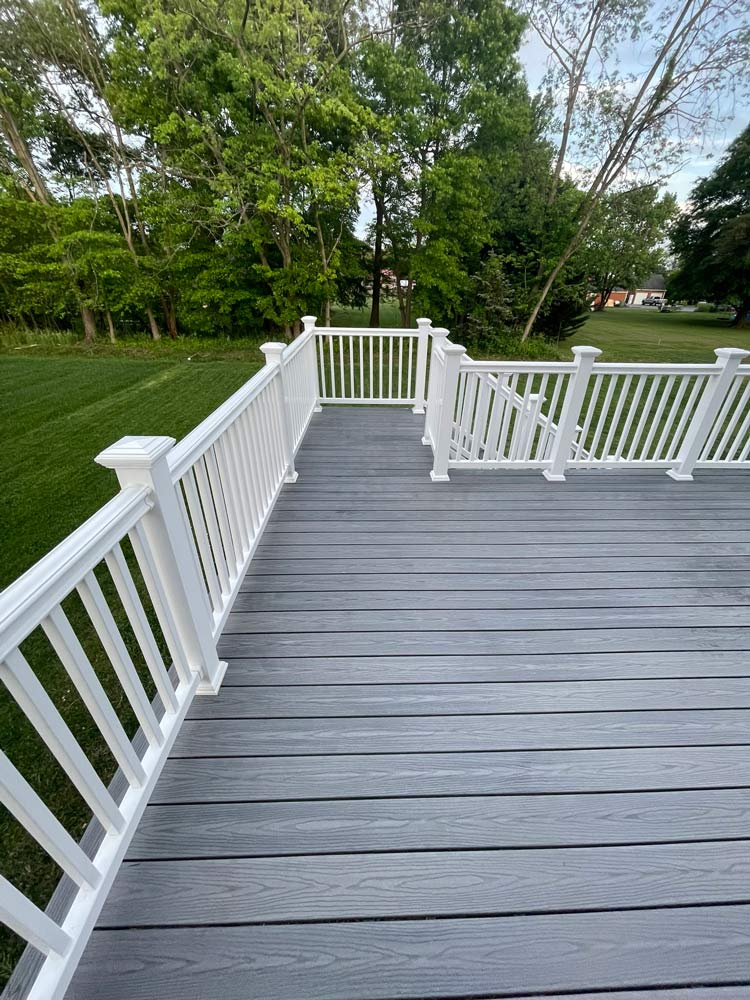 A gray composite deck with white railings overlooks a grassy yard and green trees.
