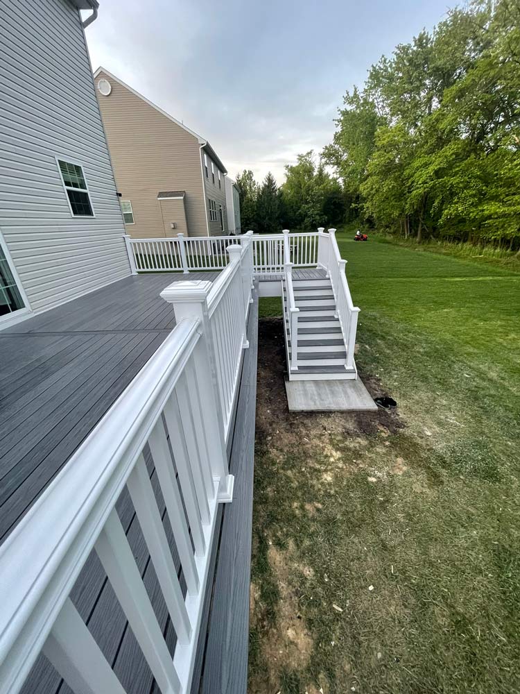 Gray deck with white railings and stairs leading to a grassy backyard next to two houses and trees.