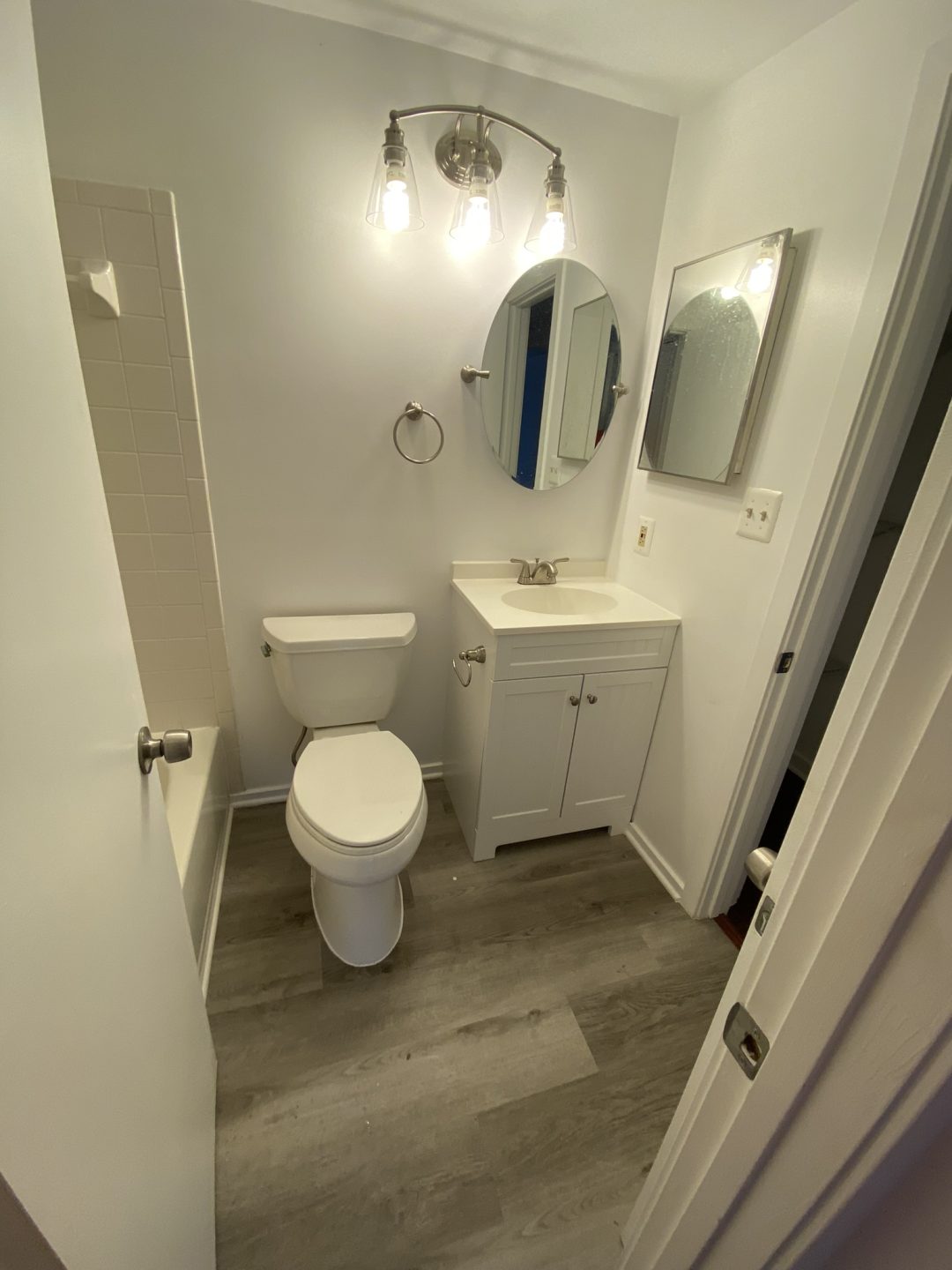 Small bathroom with a toilet, white vanity, mirror, and lights; shower stall on the left.