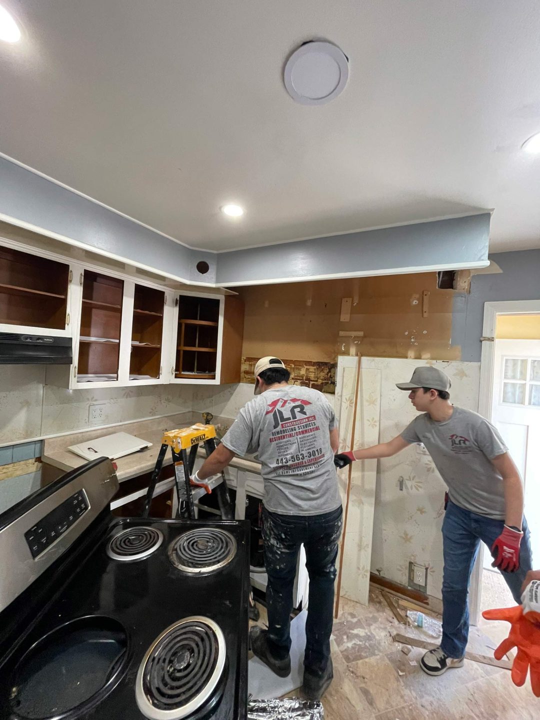 Two men in work shirts remodeling a kitchen, with cabinets open and tools and materials scattered around.