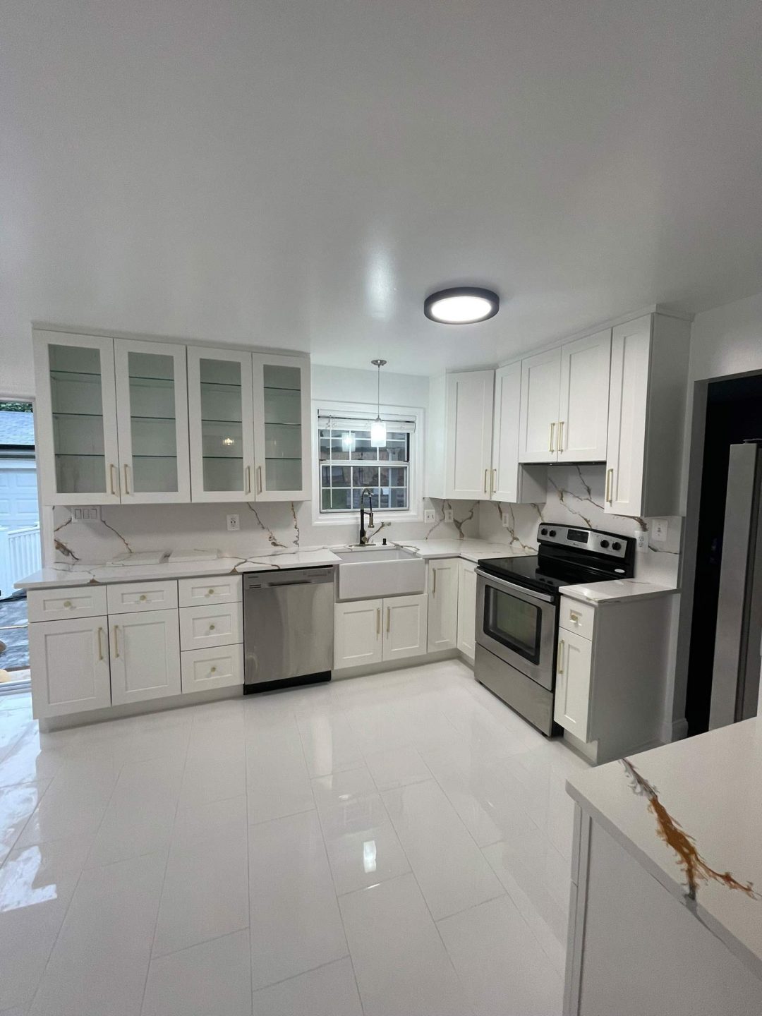 Modern white kitchen with stainless steel appliances, marble countertops, and glass-front cabinets.
