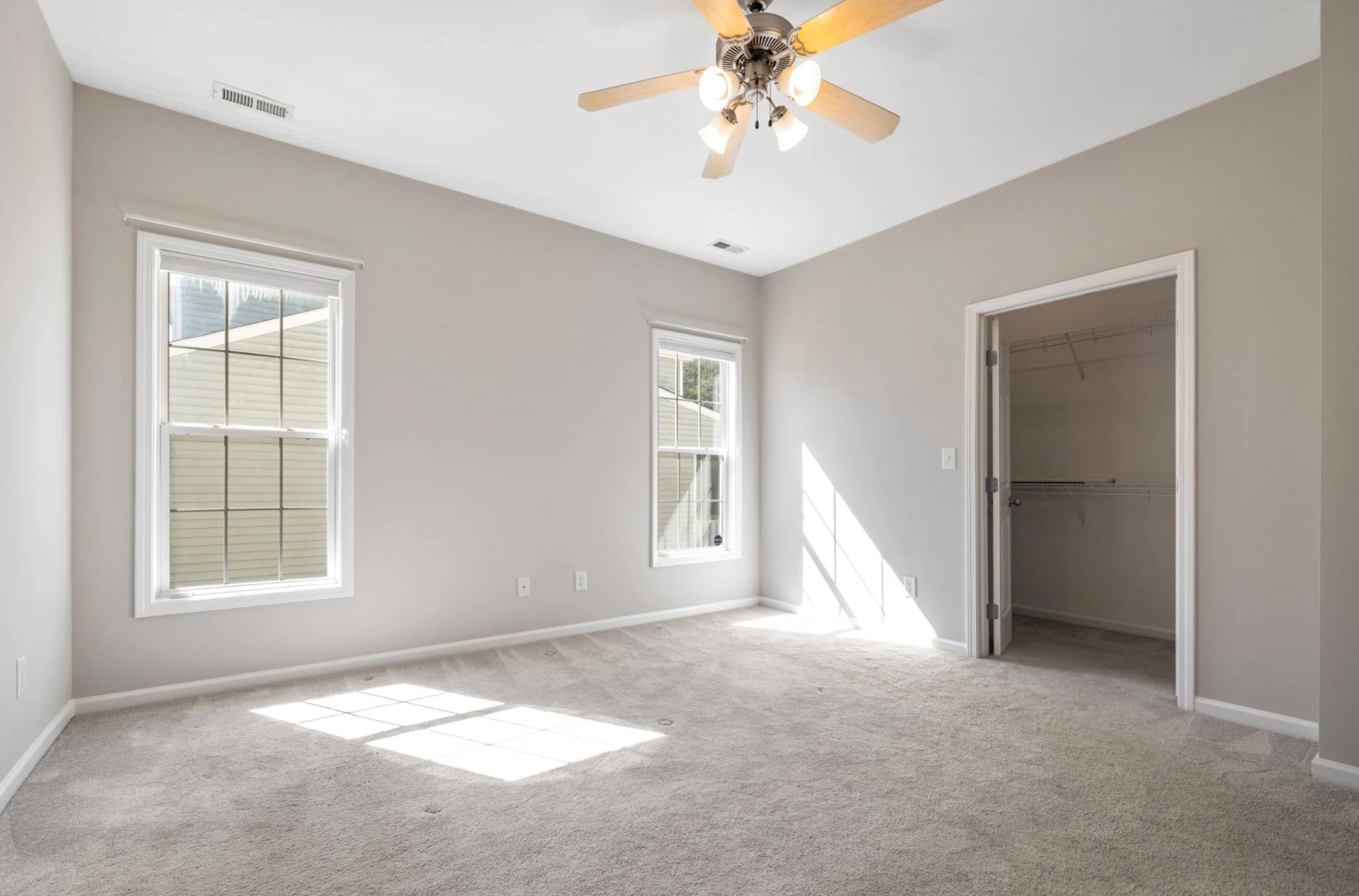 Empty bedroom with beige carpet, two windows, ceiling fan, and open walk-in closet, sunlight streaming in.