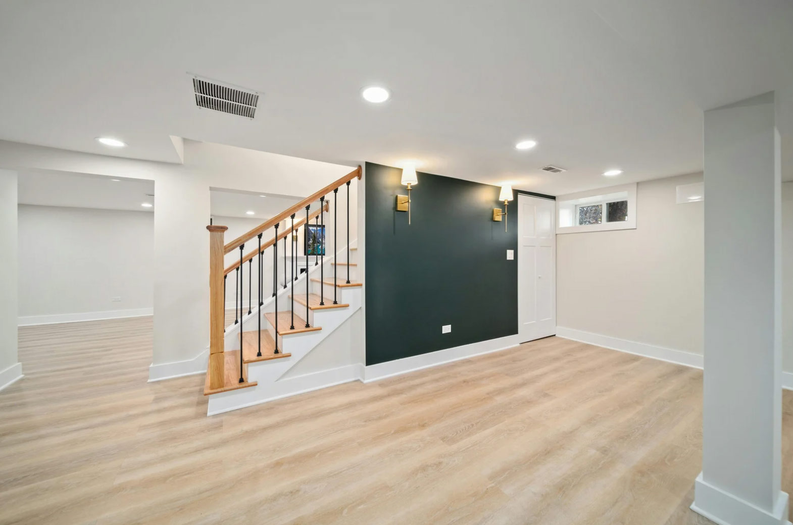 basement-3 Modern finished basement with light wood floors, white walls, a dark accent wall, and a wooden staircase.