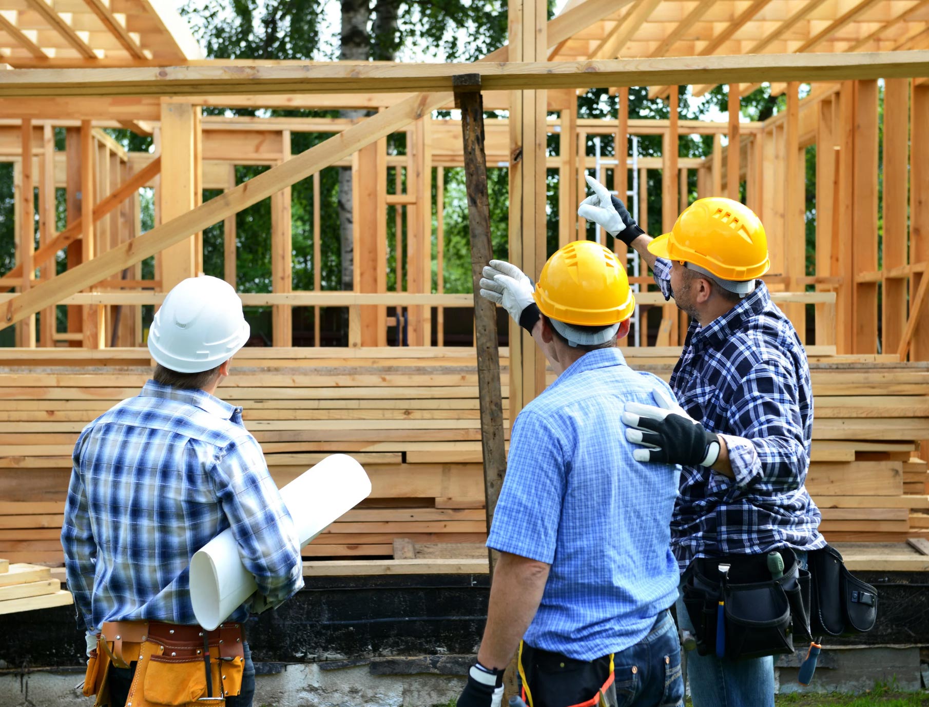 Three construction workers in hard hats discuss a wooden house frame at a building site.