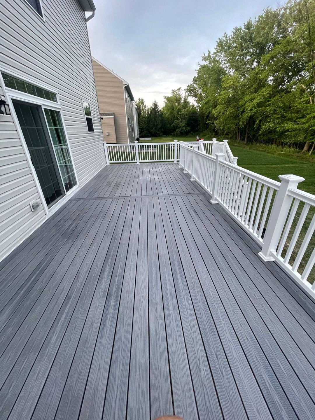 Gray wooden deck with white railing attached to a house, overlooking a green lawn and trees.