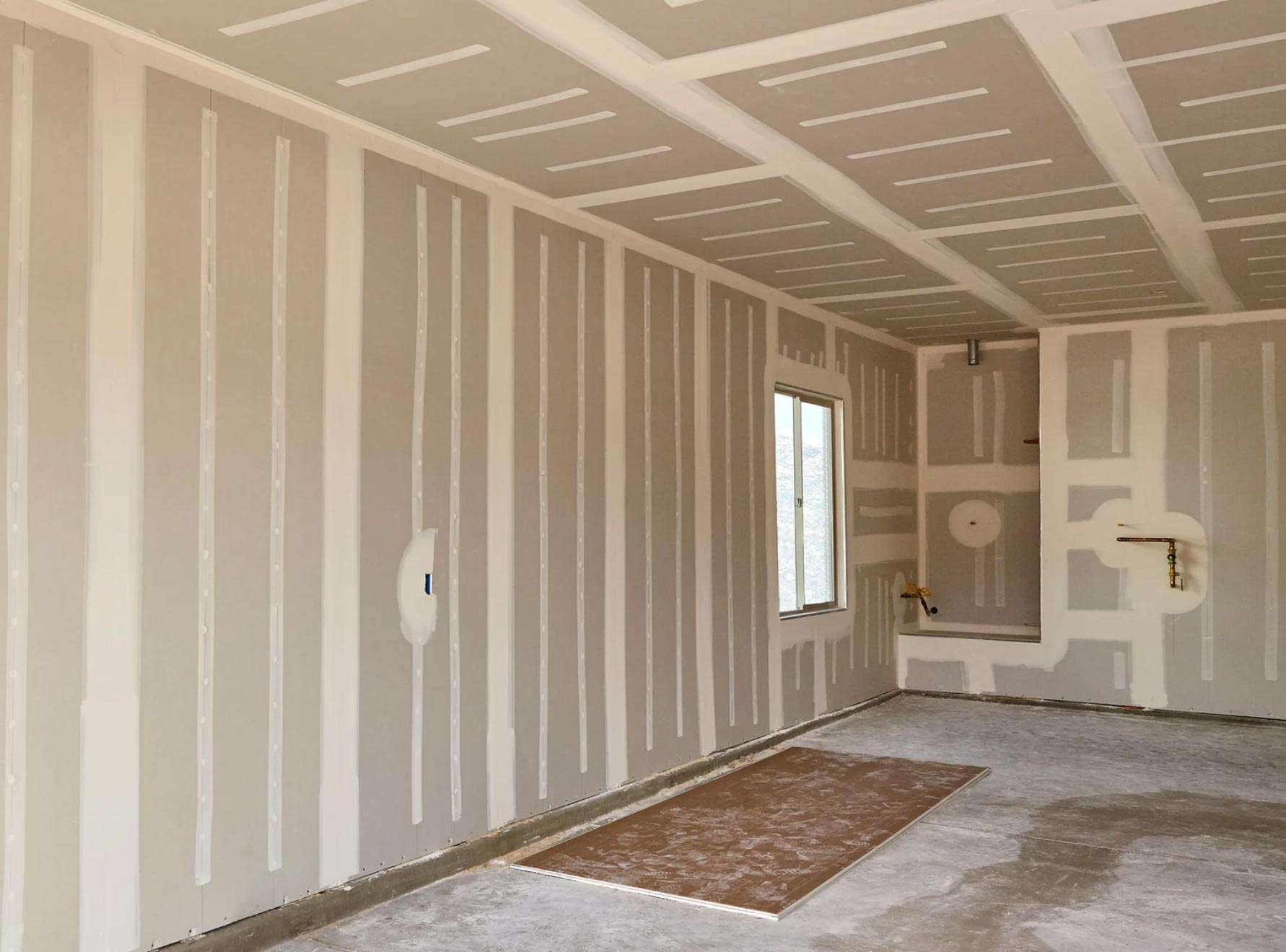 Room with unfinished drywall, taped seams, a window, and rough plumbing visible on the far wall.