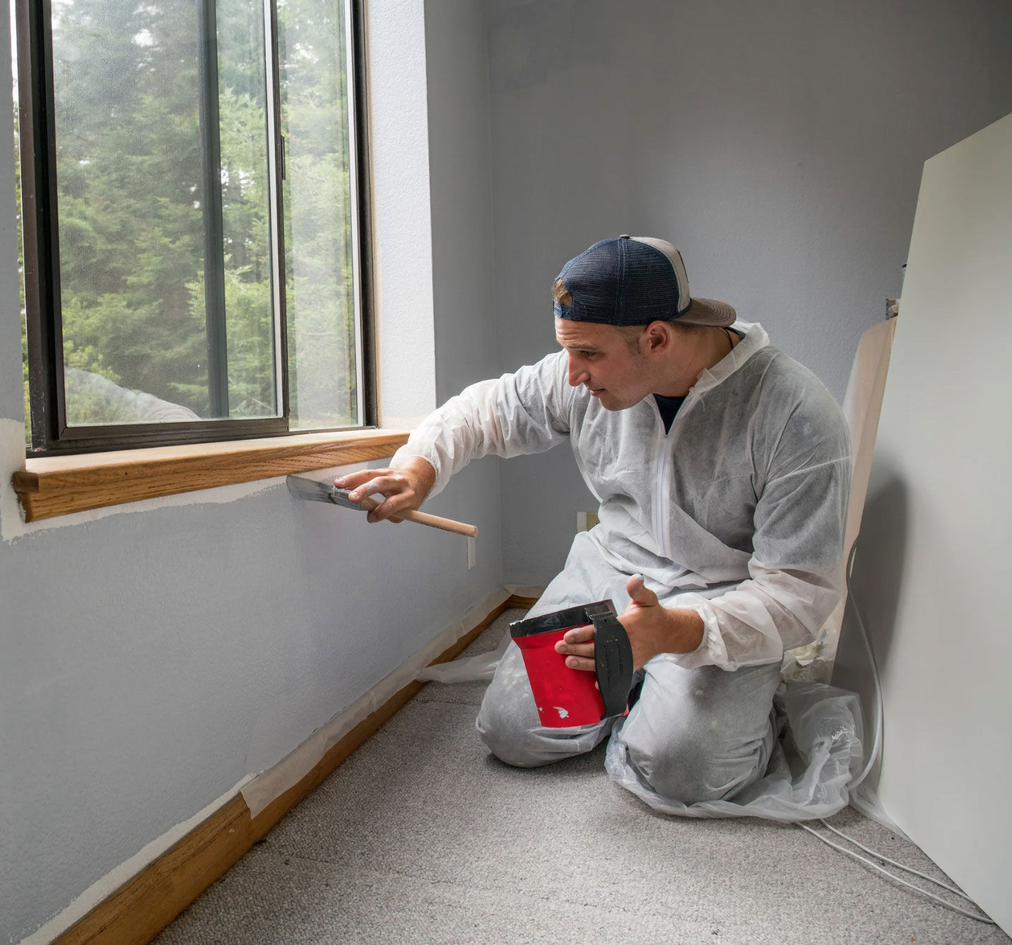 Man in coveralls painting a wall with a brush near a window, holding a paint bucket.